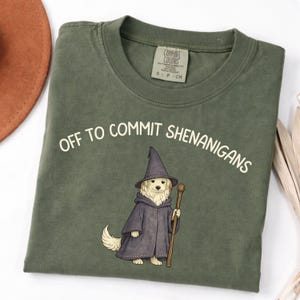 May include: An olive green t-shirt with the phrase "OFF TO COMMIT SHENANIGANS" in white text. A cartoon dog wearing a wizard hat and robe is featured below the text, holding a staff. The shirt has a Comfort Colors tag.