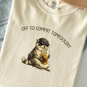 May include: Cream-coloured Comfort Colors t-shirt with a raccoon wearing sunglasses and a hat, holding a drink with a straw. The text "OFF TO COMMIT TOMFOOLERY" is arched above the raccoon illustration.