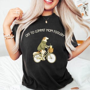 Puede incluir: Camiseta negra con el texto "OFF TO COMMIT MOM FOOLERY" y un dibujo de un perro en bicicleta. El perro lleva un suéter verde y lleva una bebida en una cesta.