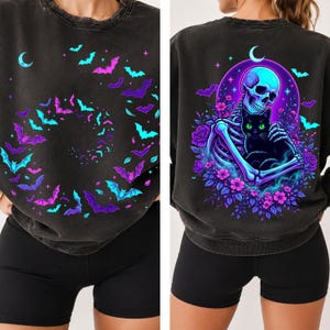 May include: A black acid-wash crewneck sweatshirt featuring a spiral pattern of neon blue and purple bats on the front. The back displays a graphic of a skeleton holding a black cat surrounded by purple flowers, a crescent moon, and bats.