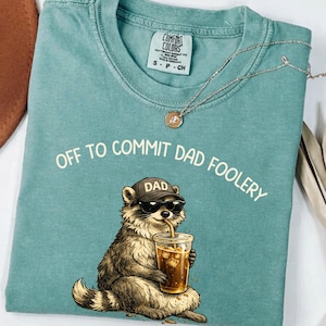 Op de afbeelding: Comfort Colors t-shirt in teal met de witte tekst "OFF TO COMMIT DAD FOOLERY". Een wasbeer met zonnebril en een "DAD" hoed is afgebeeld onder de tekst, met een drankje in zijn hand. Een zilveren ketting met een hanger ligt op het shirt.