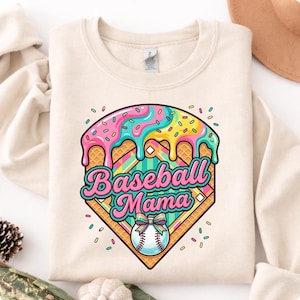 May include: Cream-colored sweatshirt featuring a colorful graphic. The graphic includes a baseball diamond design with the words "Baseball Mama" in a script font. The design incorporates ice cream-like drips and a baseball with a bow.