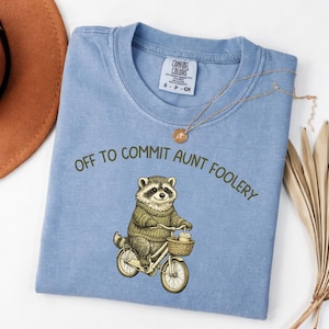 Puede incluir: Camiseta azul claro Comfort Colors con la frase "OFF TO COMMIT AUNT FOOLERY" y un mapache en bicicleta. El mapache lleva un suéter y una cesta con una taza de café. También se ven un collar dorado y un sombrero marrón.