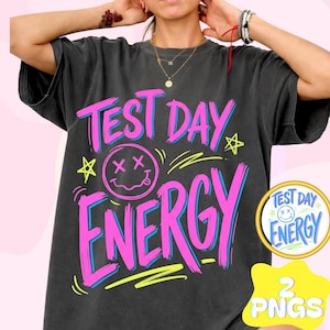 May include: Dark grey t-shirt featuring the words "TEST DAY ENERGY" in pink and blue. A smiley face with crossed-out eyes and a tongue is in the center. The shirt also has a star and yellow design.