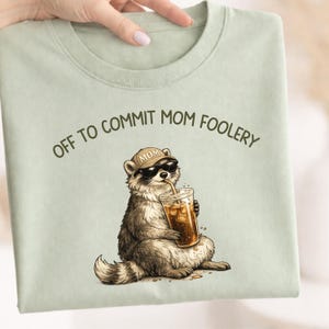 Può includere: T-shirt verde chiaro con un procione con occhiali da sole e un berretto "MOM". Il procione tiene in mano una bevanda in un bicchiere trasparente. Il testo "OFF TO COMMIT MOM FOOLERY" è arcuato sopra il procione.
