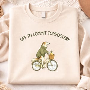 May include: Cream-colored sweatshirt with the phrase "OFF TO COMMIT TOMFOOLERY" in dark green. The graphic features a dog riding a bicycle, wearing a green sweater, and holding a drink. The bicycle has a basket.