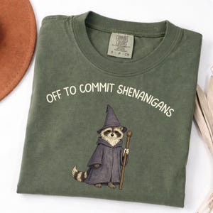 May include: Olive green t-shirt with the text "OFF TO COMMIT SHENANIGANS". The shirt features a raccoon wearing a wizard hat and robe, holding a staff. The shirt has a Comfort Colors tag.