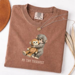 May include: A brown t-shirt with a graphic of two monkeys hugging a stuffed animal monkey. The text "MY TINY THERAPIST" is printed below the graphic. A silver necklace with a small pendant is draped on the shirt.