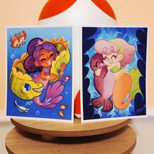 May include: Two art prints featuring cartoon mermaids and sea creatures. One shows a mermaid with purple hair and a yellow eel. The other depicts a mermaid hugging a seal. Both have a blue background.