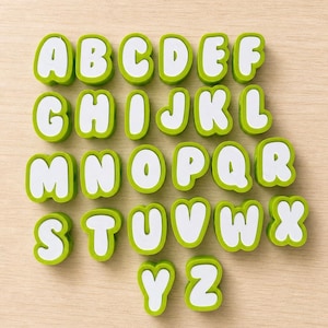 Pode incluir: Um conjunto completo de vinte e seis letras individuais do alfabeto de A a Z. Cada letra é construída com um centro branco e uma borda espessa e arredondada na cor verde-limão. As letras estão dispostas em cinco linhas ordenadas.