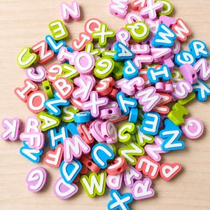 May include: A collection of small, flat, circular alphabet beads in various colors including pink, blue, green, and white. Each bead features a bold, white uppercase letter printed on the front, with a hole through the center for threading.
