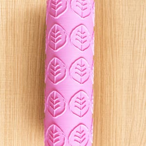 Pode incluir: Um rolo cilíndrico cor-de-rosa com um padrão em relevo repetitivo de folhas estilizadas. A superfície da ferramenta é lisa e uniforme, com os motivos de folhas dispostos numa grelha ao longo de todo o comprimento do cilindro.