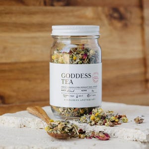 May include: A clear glass jar of Goddess Tea with a white lid and label. The label lists ingredients like chamomile and rose. A wooden spoon holds tea leaves, and loose tea is scattered around. The jar is 8 oz.
