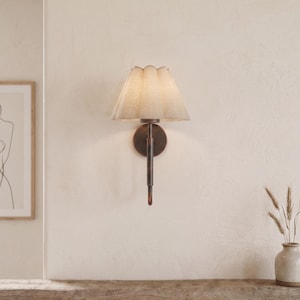 May include: A wall-mounted light fitting with a scalloped fabric lampshade. The fixture has a dark bronze finish and a long, slender arm. The shade is a light beige colour, and the sconce is mounted on a cream-coloured wall.