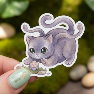 May include: A sticker featuring a cartoon cat with large green eyes and a purple body. The cat is playing with a twenty-sided die. The sticker has a white border and is held by a person.