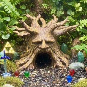 Può includere: Un terrario a tema fantasy con una dettagliata struttura marrone a forma di albero con un volto, circondata da vegetazione. Sono presenti anche una spada in miniatura, gemme colorate e una pianta con la bocca aperta.