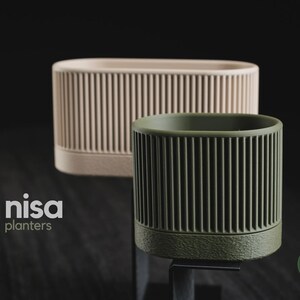 Op de afbeelding: Twee cilindrische plantenbakken met verticaal geribbelde texturen. De ene plantenbak is lichtbeige en de kleinere op de voorgrond is olijfgroen. De tekst nisa planters en h3liø design is zichtbaar op de afbeelding.
