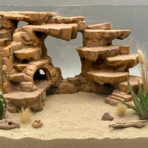 Könnte beinhalten: Braune, gestufte Felsformation mit Höhle und Treppen, entworfen für ein Terrarium oder Aquarium. Die Szene umfasst Sand, Kakteen und getrocknete Pflanzen, wodurch eine wüstenähnliche Umgebung entsteht. Die Struktur ist etwa 30 cm breit.