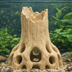 May include: A beige, tree stump-shaped aquarium decoration with multiple holes and a hollow center. The decoration sits on a bed of small stones in a glass aquarium, with green plants and a fish visible in the background.