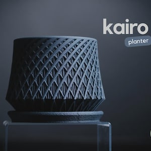 Peut inclure: Un cache-pot Kairo gris foncé avec un motif en treillis diagonal répétitif sur sa surface extérieure. Le cache-pot repose sur une soucoupe circulaire assortie. Le texte kairo planter est visible en haut à droite, et un petit logo h3liø design est en bas à droite.