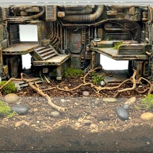 May include: A miniature diorama depicting a futuristic, industrial interior. The structure is weathered with brown, grey, and green tones, featuring platforms, stairs, and exposed pipes. The base includes soil, rocks, moss, and twigs, creating a realistic, post-apocalyptic scene.