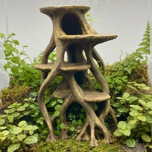 May include: A tree-shaped terrarium ornament with multiple platforms and a hollowed-out centre. The ornament is surrounded by green plants and moss, creating a naturalistic look. The colour palette includes shades of brown and green, evoking an earthy feel.