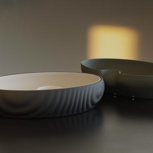 Pu&ograve; includere: Due moderne ciotole ovali in ceramica. Una &egrave; grigio scuro con un motivo testurizzato a forma di onda all'esterno e un interno bianco. L'altra &egrave; verde oliva con una finitura liscia. Entrambe sono su una superficie scura.