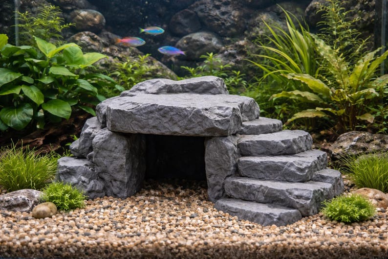 May include: A gray, stone-like aquarium decoration featuring a cave and steps. The structure is surrounded by green plants, gravel, and colorful fish. This decorative element adds a naturalistic aesthetic to any aquarium.