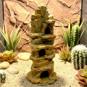 May include: A decorative, layered rock structure with multiple openings, designed for a terrarium or aquarium. The structure is tan and brown, surrounded by sand, rocks, and various succulents and cacti. The background features a textured rock wall.