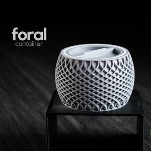 Puede incluir: Un recipiente gris de forma esférica con un complejo patrón geométrico de celosía abierta en el exterior. La parte superior presenta una tapa circular plana con un asa central. El texto "foral container" y "h3liø design" aparecen en la imagen.