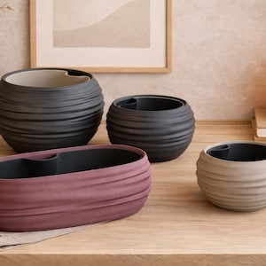 Peut inclure: Une collection de jardinières décoratives en céramique de différentes tailles et couleurs. Les jardinières sont dans des tons de gris, mauve et beige, avec un design texturé et nervuré. L'arrangement comprend un grand vase avec des branches séchées et une lanterne lumineuse.