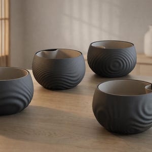 Peut inclure: Quatre bols en céramique gris foncé avec un motif texturé en forme de vagues à l'extérieur et un intérieur beige clair. Chaque bol a une petite encoche sur le bord. Les bols sont disposés sur une surface en bois clair.