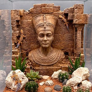 Peut inclure: Un terrarium sur le thème égyptien, avec un buste de pharaon et des ruines en fausse pierre. Plusieurs petits lézards avec des marques orange, noires et blanches sont visibles. Le terrarium comprend également des cactus, des succulentes et de petits rochers.