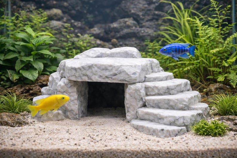 May include: A decorative aquarium ornament resembling a stone structure with steps and an archway. A yellow and a blue fish swim near the ornament. Green plants and sand complete the underwater scene.