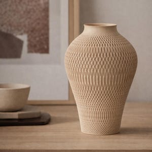 Pode incluir: Um vaso decorativo bege com um design de treliça. O vaso tem uma base larga que se estreita para um pescoço estreito. A superfície tem uma aparência texturizada e tecida. O vaso está em uma mesa de madeira, com uma tigela e uma obra de arte no fundo.