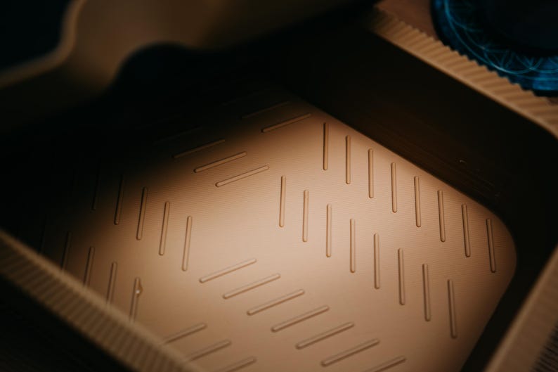 May include: A close-up shot of a tan, rectangular container interior. The base features a textured pattern of diagonal, parallel lines. The lighting creates shadows, highlighting the texture and shape of the container.