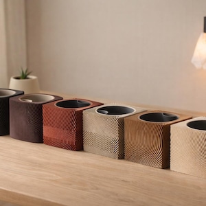 Op de afbeelding: Zes vierkante plantenbakken met getextureerde, golvende patronen op een rij. De plantenbakken zijn in tinten zwart, bruin en beige, met een donker interieur. Een kleine plant in een witte pot staat op de achtergrond.