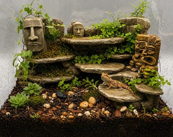 May include: A terrarium with a bearded dragon lizard, featuring stone sculptures of faces and tiered platforms. The habitat includes live plants, moss, and small rocks, creating a naturalistic environment. The glass enclosure provides a clear view of the setup.
