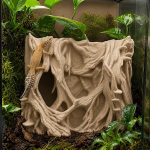Puede incluir: Un escondite para reptiles beige con un diseño texturizado en forma de árbol, con un gecko. El escondite tiene múltiples aberturas y está rodeado de vegetación, creando un entorno de terrario natural.