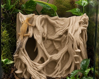 May include: A beige reptile hide with a textured, tree-like design, featuring a gecko. The hide has multiple openings and is surrounded by greenery, including plants and moss, creating a naturalistic terrarium environment.