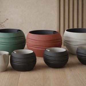 Puede incluir: Colección de macetas de cerámica en varios tamaños y colores, incluyendo verde, terracota y blanco. Las macetas tienen un diseño texturizado en forma de onda y están dispuestas sobre una superficie de madera. Interiores oscuros.