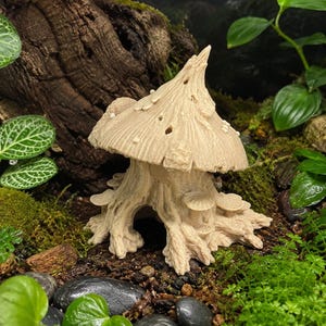 May include: A beige mushroom sculpture with a textured cap and stem, featuring small holes and attached mushroom-like details. The sculpture is set amidst green moss, small rocks, and foliage, creating a miniature garden scene.