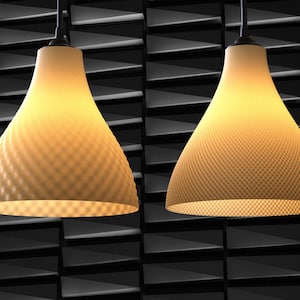 Op de afbeelding: Twee hanglampen met getextureerde, crèmekleurige kappen hangen tegen een donkere, geometrische achtergrond. De kappen hebben een uitlopende, klokvormige vorm en geven een warme gloed. De lampen zijn opgehangen aan zwarte snoeren.