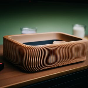 May include: A wooden, rectangular box with a unique, ridged design. The box is light brown with a dark interior and sits on a wooden surface. A black remote control is in the foreground. The box appears to be a modern home decor item.
