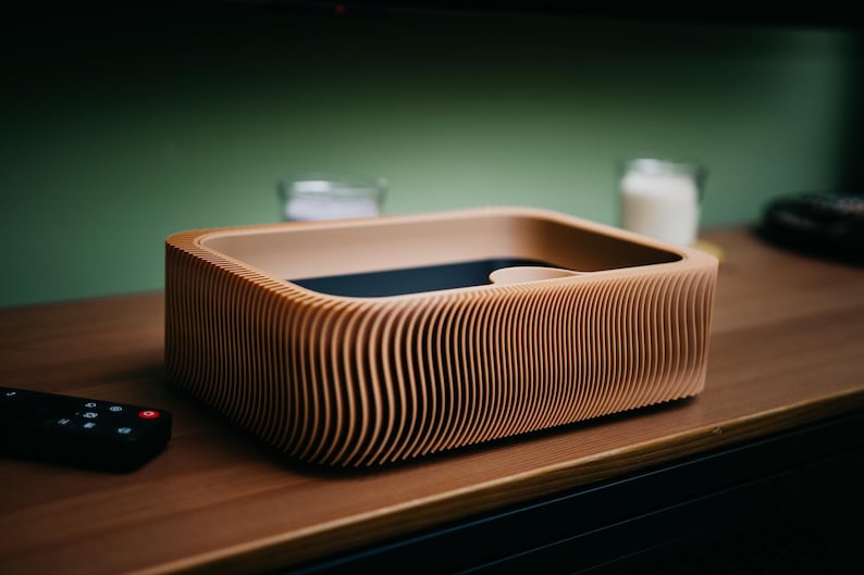 May include: A wooden, rectangular box with a unique, ridged design. The box is light brown with a dark interior and sits on a wooden surface. A black remote control is in the foreground. The box appears to be a modern home decor item.