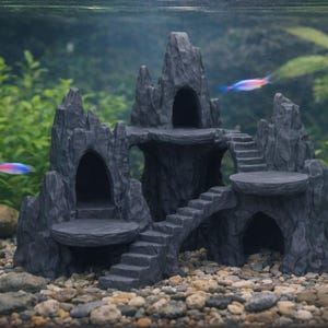 May include: A dark grey aquarium castle ornament with multiple levels, stairs, and arched openings. The castle is surrounded by gravel and aquatic plants, with two colourful fish swimming nearby.