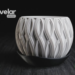 Puede incluir: Una maceta esférica blanca con un patrón geométrico texturizado de líneas verticales curvas y formas de diamante. La maceta se asienta sobre un plato negro curvado. El texto "velar planter" aparece en la esquina superior izquierda.