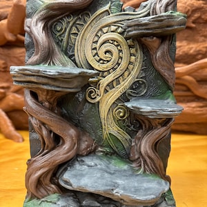 Peut inclure: Un présentoir vertical composé de trois plateformes plates en pierre, soutenues par des structures brunes texturées rappelant des arbres. Le panneau central présente un relief en spirale doré sur un fond vert foncé et noir.