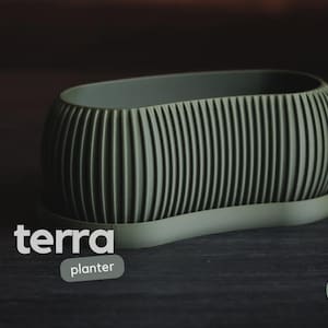 May include: An oval-shaped sage green planter with a ribbed, vertical texture on the exterior. The planter sits on a matching oval saucer. The text "terra planter" appears in white on the left, and a circular "h3liø design" logo is on the right.