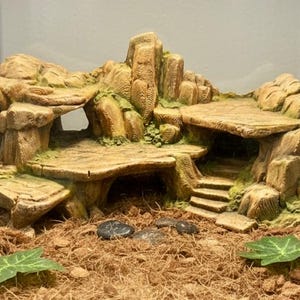 May include: A detailed, multi-level terrarium decoration in shades of brown and tan, resembling a rocky landscape with platforms, arches, and steps. Green moss accents add a touch of colour. Ideal for reptile habitats.